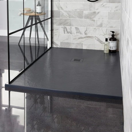 A dark charcoal gray slate-look shower traywith a built-in drain, installed in a modern walk-in shower with gray marble-look tiles and a black-framed glass door.