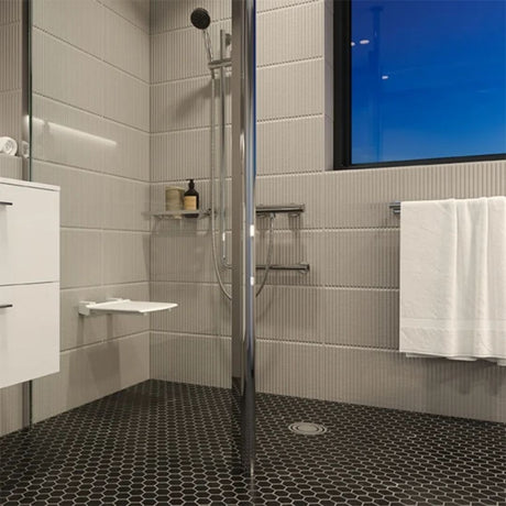 Modern walk-in shower with a clear glass door, chrome shower fixture, black hexagonal patterened vinyl floor, white wall tiles, a white towel, and a blue-tinted window.