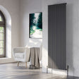 A tall black radiator with vertical fluted panels, mounted on a textured white wall in a modern living room setting next to a white armchair and a framed forest print.