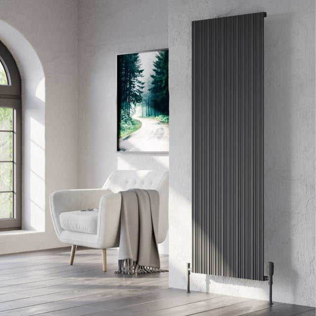A tall black radiator with vertical fluted panels, mounted on a textured white wall in a modern living room setting next to a white armchair and a framed forest print.