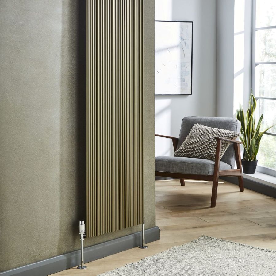 A tall, narrow ribbed radiator in a warm bronze finish, mounted on a textured gray wall in a modern living room setting with a wooden armchair and window in the background.